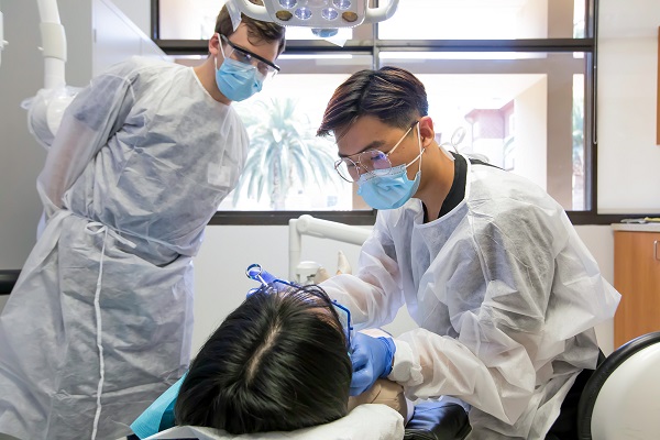 A man watching a dentist who is treating a patient at a small dental clinic.