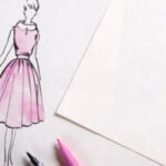 A sketch of a model in a designer dress is drawn on a sheet pf paper; some papers, pins and coloured pens are seen on the table too