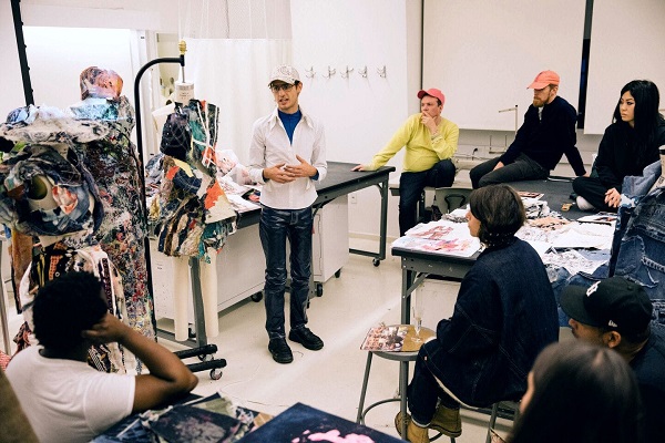 An instructor is teaching, students are listening and there are lots of drawings kept on the table and fabric pieces draping the mannequin on the side